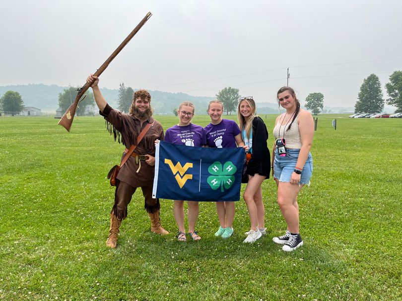WVU Mountaineer mascot poses with four girls holding a flag with the flying WV and 4-H clover on it.