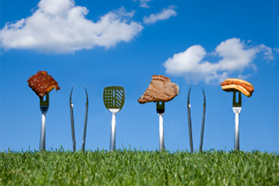 fun photo of bbq instruments standing vertically in grass with blue sky behind, some items have food on them