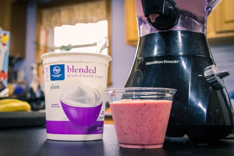 Glass of smoothie with yougurt container and blender in kitchen. 