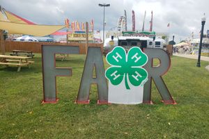 Metal FAIR sign. Instead of the letter I, there is a green 4-H clover. Carnival tents and rides are in the background of the image.