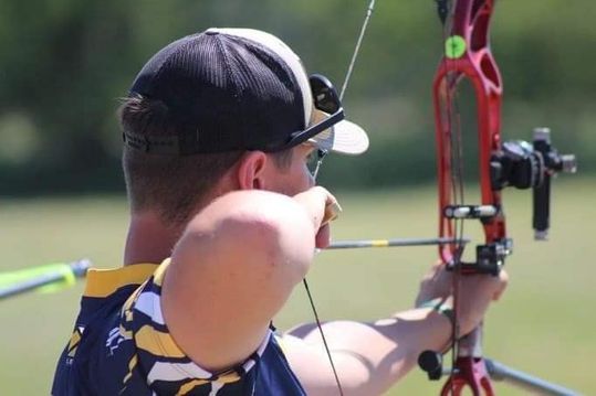 A young man in a gold/blue shirt and baseball cap pulls the bow back as he prepares to hit his archery target.