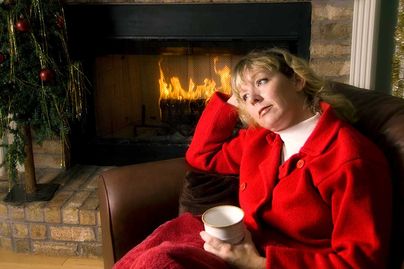 A woman sits by the fireplace with mug in hand looking as if she is in thought.