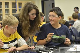 Two youth boys in safety glasses work on a computer project, as a female teacher leans over to look at their work at 4-H code camp