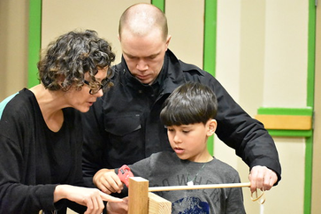 A boy using a hot glue gun to assemble a wooden building kit with assistance from a man and woman