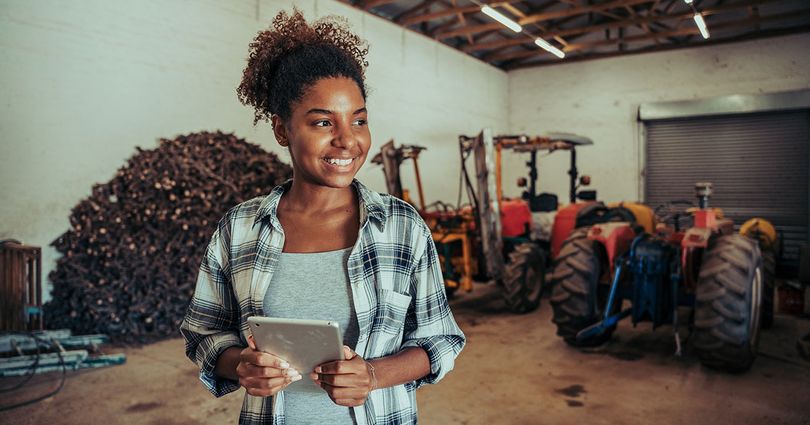 A woman holds a tablet standing in a barn with tractors in the background.