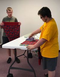 A youth ironing a pillowcase. A second youth watching and holding a pillowcase.