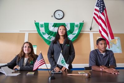 Two teens sitting at a table and one teen standing with 4-H and American flags on table and in the background.