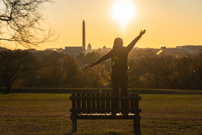 A woman stands on a bench lifting her hand toward the sun with the Washington DC Capitol skyline in the horizon.