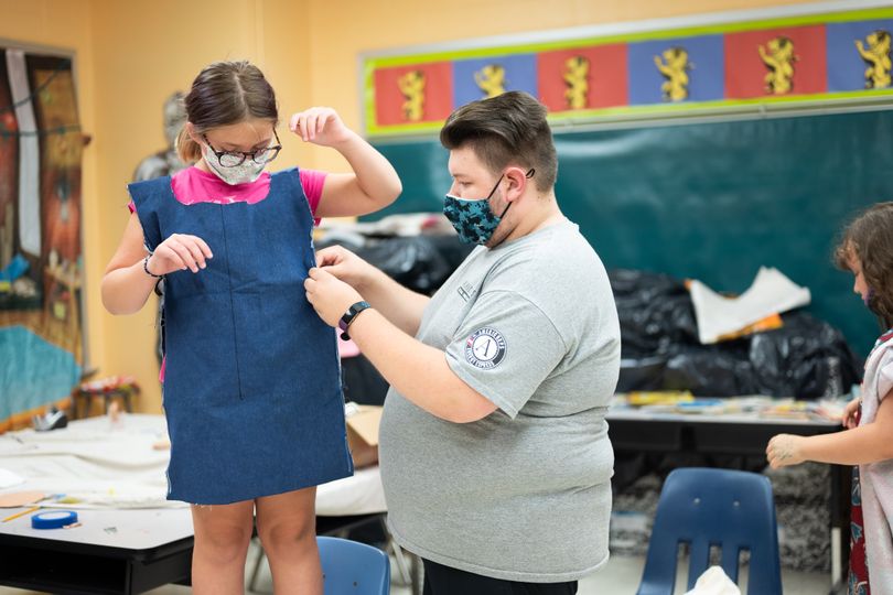 Little girl holds arms up as a man pins fabric around her to make into a dress.