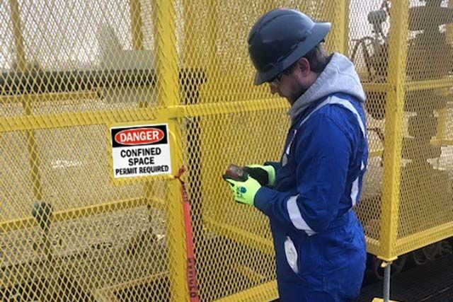An oil and gas worker dressed in blue safety attire and hard hat considers entering a permit-required confined space.