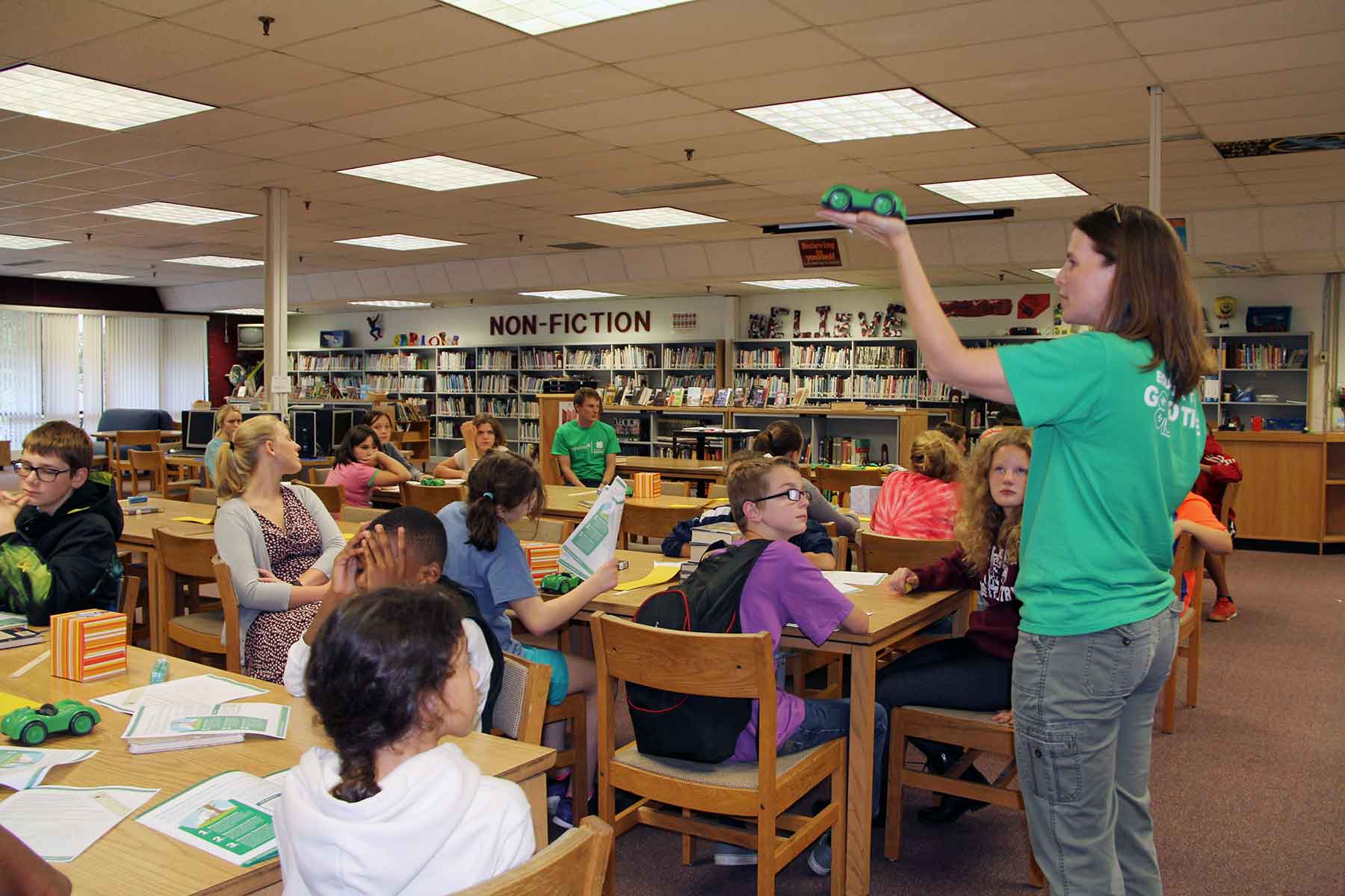 Jen Robertson-Honecker demonstrates STEM principles with a race car, showing students seated in a library.